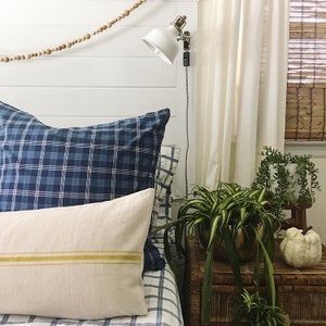 May include: A bedroom with a white headboard, blue and white plaid bedding, a white lamp with a gold base, and a plant in a brown pot on a wicker nightstand.