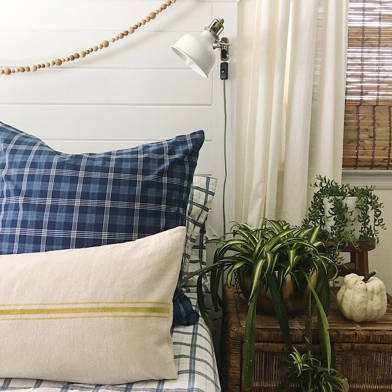 May include: A bedroom with a white headboard, blue and white plaid bedding, a white lamp with a gold base, and a plant in a brown pot on a wicker nightstand.