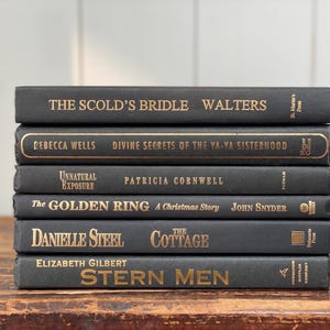 May include: A stack of six hardcover books with dark gray covers and gold lettering. Titles include "The Scold's Bridle," "Divine Secrets of the Ya-Ya Sisterhood," and "Stern Men." The books are arranged on a wooden surface.