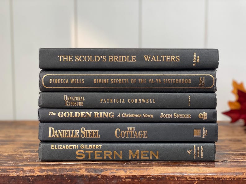 May include: A stack of six hardcover books with dark gray covers and gold lettering. Titles include "The Scold's Bridle," "Divine Secrets of the Ya-Ya Sisterhood," and "Stern Men." The books are arranged on a wooden surface.