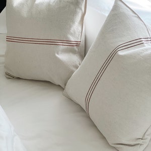 May include: Two square off-white throw pillows made of a textured fabric. Each pillow features two thin, parallel horizontal stripes in a dark reddish-brown color running across the front surface. The pillows are positioned on a white surface.