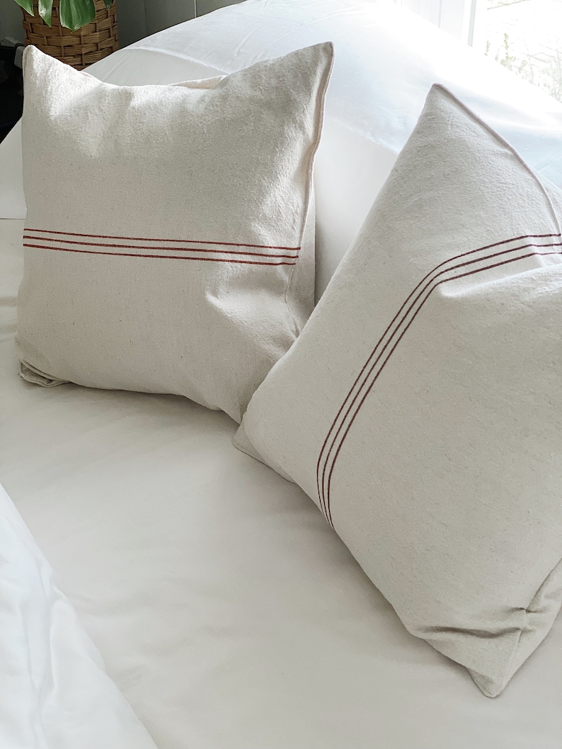 May include: Two square off-white throw pillows made of a textured fabric. Each pillow features two thin, parallel horizontal stripes in a dark reddish-brown color running across the front surface. The pillows are positioned on a white surface.