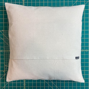 May include: A square, off-white throw pillow cover featuring a plain, solid-colored fabric texture. The cover includes a horizontal seam across the lower portion and a small, dark rectangular brand label attached to the right side seam.