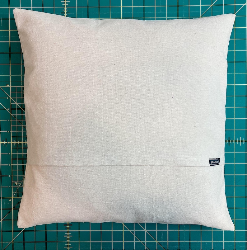 May include: A square, off-white throw pillow cover featuring a plain, solid-colored fabric texture. The cover includes a horizontal seam across the lower portion and a small, dark rectangular brand label attached to the right side seam.