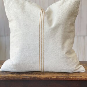 May include: A square off-white throw pillow made of a textured fabric. The pillow features a vertical design element consisting of three thin, parallel golden-brown stripes running down the center of the front face of the cushion cover.