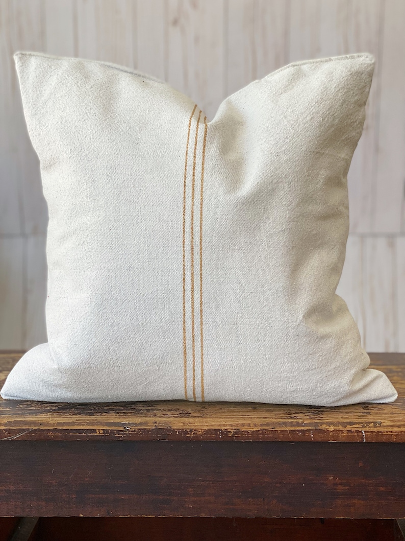 May include: A square off-white throw pillow made of a textured fabric. The pillow features a vertical design element consisting of three thin, parallel golden-brown stripes running down the center of the front face of the cushion cover.
