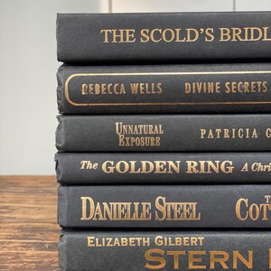 May include: A stack of six hardback books with dark gray covers and gold lettering. Titles include "The Scold's Bridle," "Divine Secrets," and "Stern Men." The books are arranged on a wooden surface.