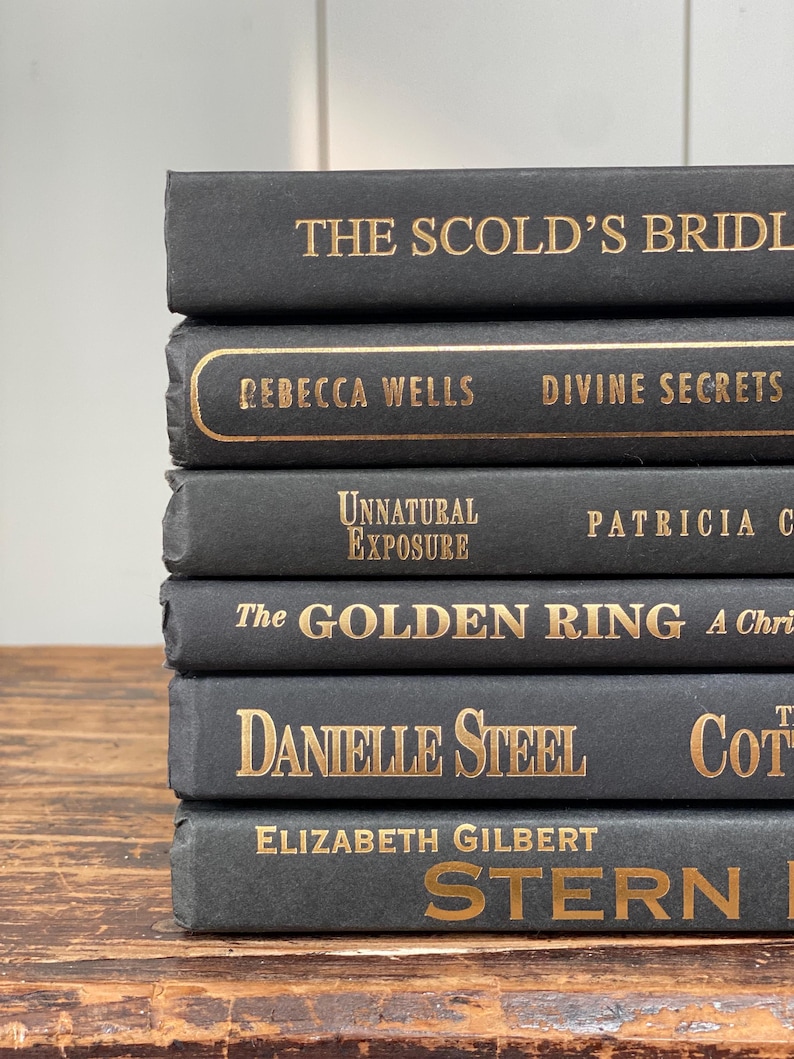 May include: A stack of six hardback books with dark gray covers and gold lettering. Titles include "The Scold's Bridle," "Divine Secrets," and "Stern Men." The books are arranged on a wooden surface.