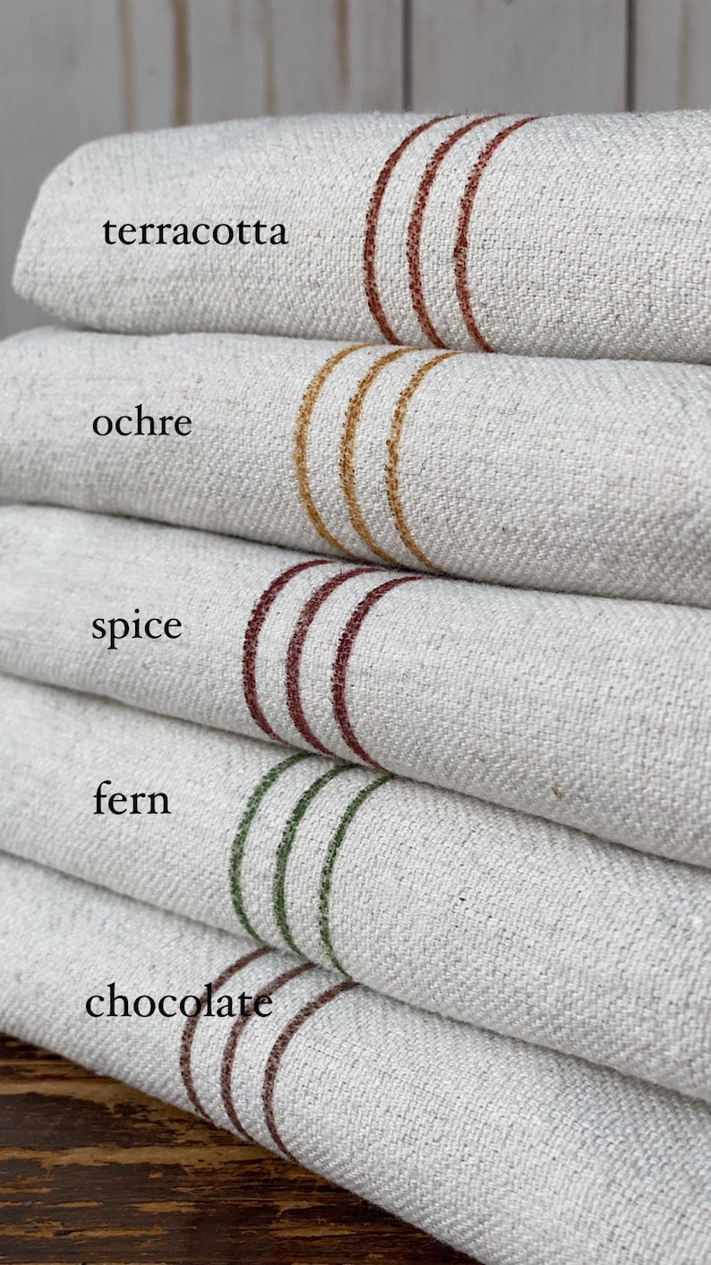 May include: A stack of five off-white textured fabric towels, each featuring three horizontal stripes in different colors. The colors are labeled as terracotta, ochre, spice, fern, and chocolate, with each label placed next to its corresponding stripe.