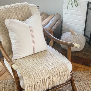 May include: A wooden armchair with brass-capped armrests features a cream-colored throw blanket with a textured weave and fringe, topped with a square off-white pillow accented by a single vertical red stripe running down the center of the fabric.