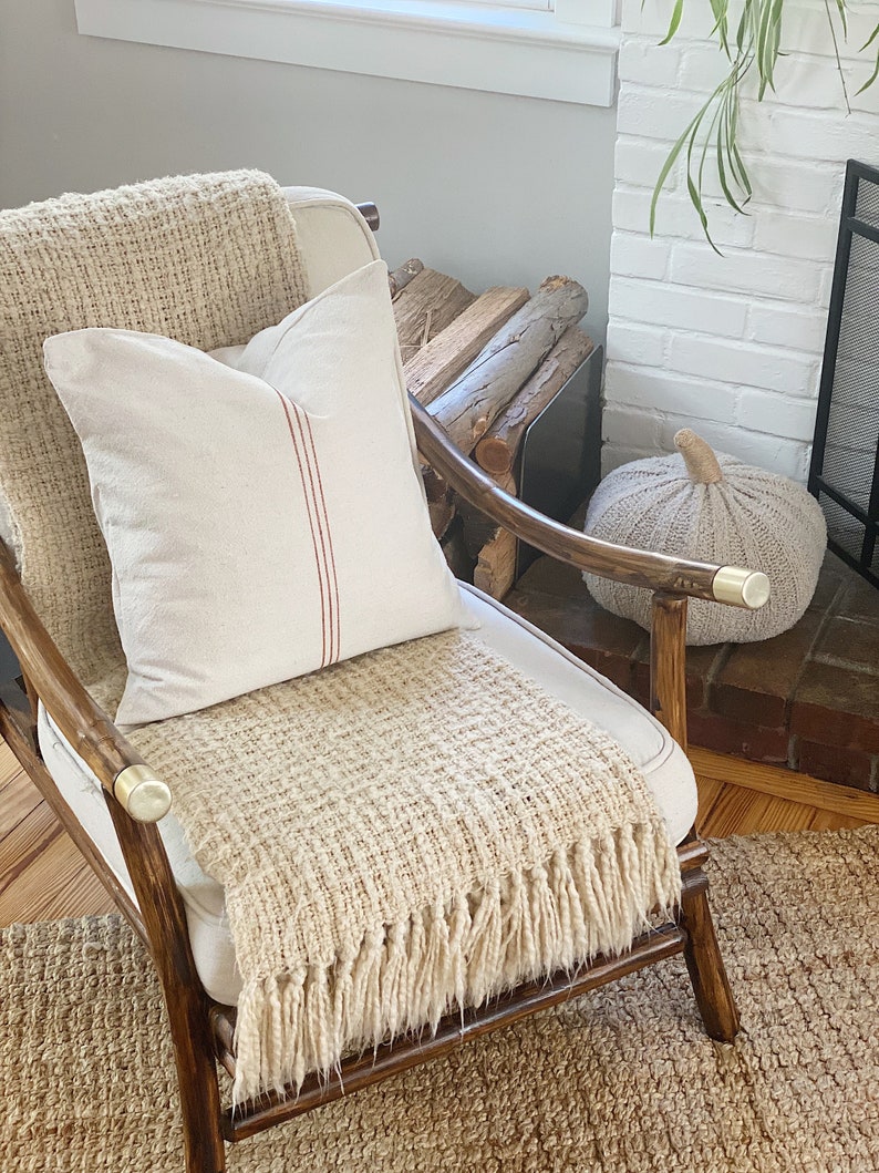 May include: A wooden armchair with brass-capped armrests features a cream-colored throw blanket with a textured weave and fringe, topped with a square off-white pillow accented by a single vertical red stripe running down the center of the fabric.