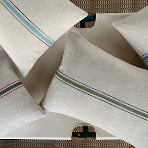 May include: Four decorative throw pillows with different colored stripes. The pillows are made of a light beige linen fabric and have a subtle texture. The stripes are in shades of blue, green, red, and brown.