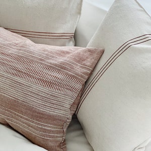 May include: Three rectangular and square throw pillows featuring a textured fabric. One pillow is a deep terracotta color with white horizontal stripes and a woven pattern, while the other two are off-white with thin, dark red horizontal stripes.