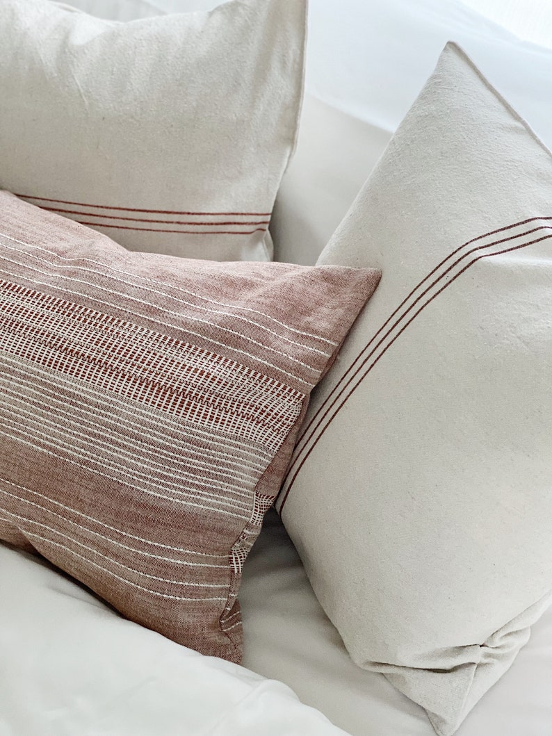 May include: Three rectangular and square throw pillows featuring a textured fabric. One pillow is a deep terracotta color with white horizontal stripes and a woven pattern, while the other two are off-white with thin, dark red horizontal stripes.