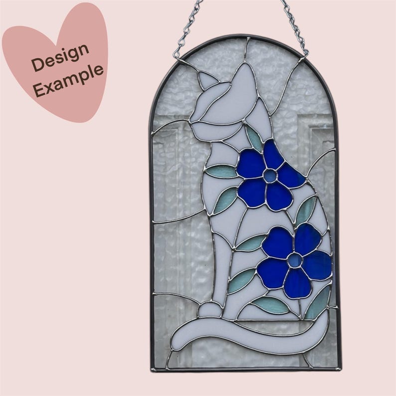May include: A stained glass window hanging with a white cat design with blue flowers. The cat is sitting with its tail curled around its legs.