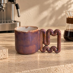 Peut inclure: Une tasse en céramique avec une finition brillante irisée violette et brune. La tasse présente une poignée unique, épaisse, ondulée et sinueuse qui forme une forme rectangulaire. L'intérieur de la tasse est blanc et le bord est arrondi.
