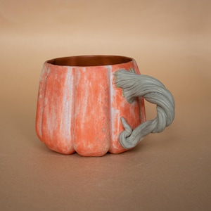 May include: Orange ceramic mug shaped like a pumpkin with a gray, twisted handle. The mug has a textured surface with white accents. The interior is brown.