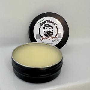 May include: A black tin with a white label that says "Brothers' Spiced Tobacco Beard Balm" with a cartoon image of a man with a beard. The tin is open and the balm is visible inside.