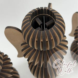 May include: A three-dimensional angel figurine constructed from layered, laser-cut wood pieces with dark brown edges and light tan faces. The design features a circular head, flared body, and wing-like projections. The text Colorful Walk is visible.
