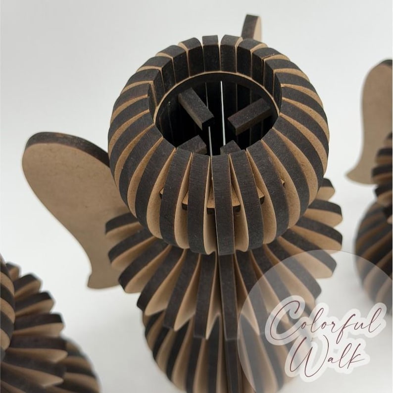 May include: A three-dimensional angel figurine constructed from layered, laser-cut wood pieces with dark brown edges and light tan faces. The design features a circular head, flared body, and wing-like projections. The text Colorful Walk is visible.