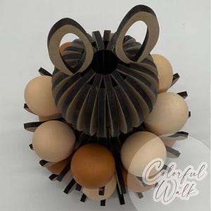 Puede incluir: Un portahuevos decorativo con un dise&ntilde;o de papel negro y beige, de forma circular y con recortes en forma de orejas. Contiene varios huevos marrones y beige. El texto "Colorful Walk" es visible.