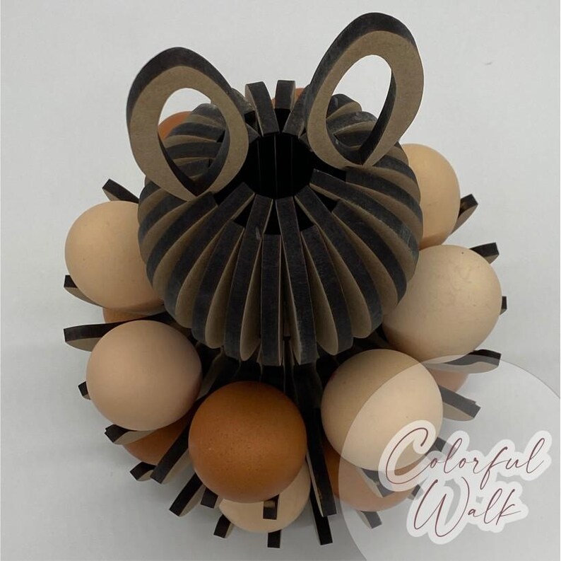 Puede incluir: Un portahuevos decorativo con un dise&ntilde;o de papel negro y beige, de forma circular y con recortes en forma de orejas. Contiene varios huevos marrones y beige. El texto "Colorful Walk" es visible.