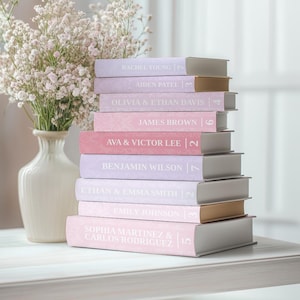May include: A collection of personalised books in shades of pink and lavender. Each book displays a name and a number. A white vase with white flowers is in the background. The books are stacked on a white surface.
