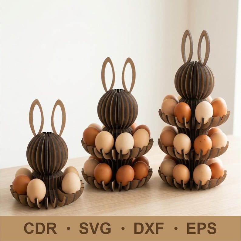 Puede incluir: Tres soportes de madera marr&oacute;n para huevos de Pascua con forma de conejito, cada uno con niveles para huevos. Los soportes est&aacute;n llenos de huevos de varios colores. El texto "CDR &bull; SVG &bull; DXF &bull; EPS" est&aacute; en la parte inferior.