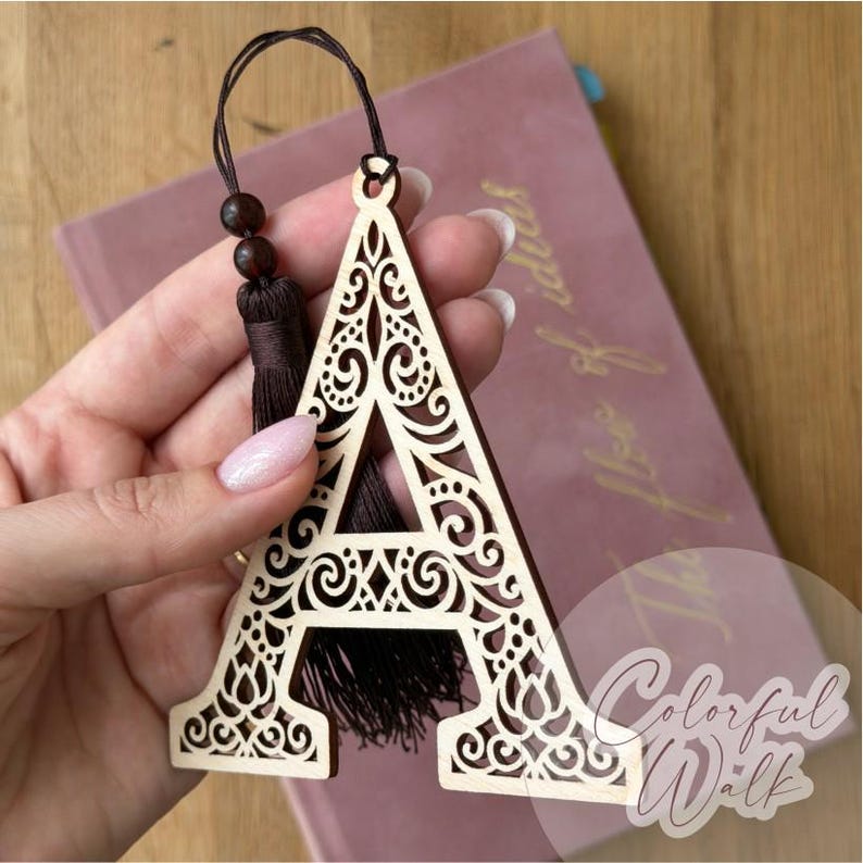 May include: A light-colored wooden letter A ornament featuring intricate laser-cut scrollwork patterns. The letter includes a dark brown cord loop with two small wooden beads and a matching dark brown fabric tassel attached to the top.