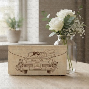 May include: A wooden wedding keepsake box with a laser-etched design of a vintage car carrying a bride and groom. The car has the words "Sarah + Caleb" and "Just Married" on it. The box is light brown and is next to a vase of white roses.
