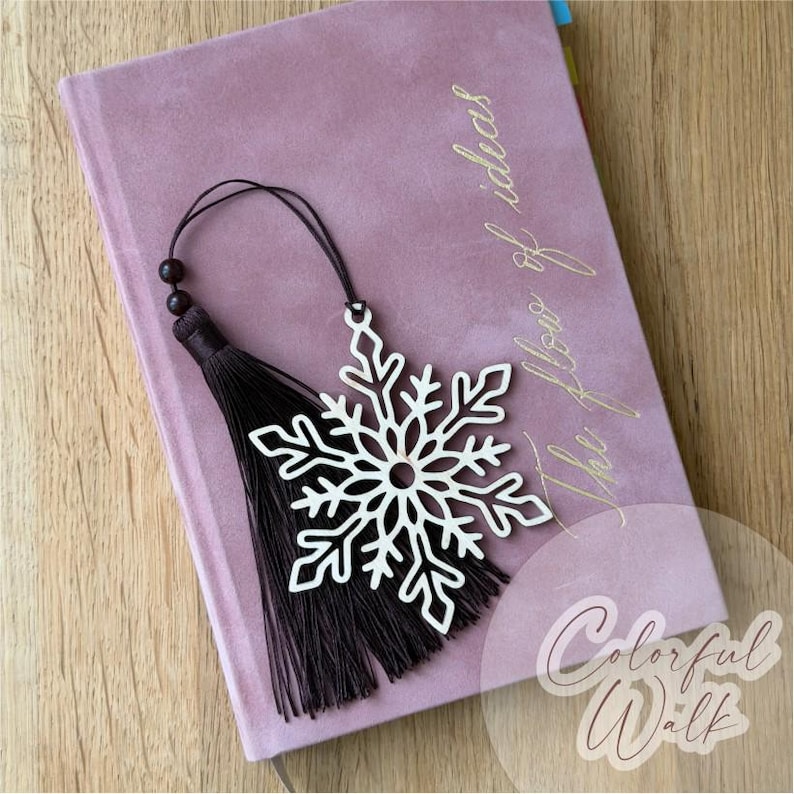 K&ouml;nnte beinhalten: Ein wei&szlig;es schneeflockenf&ouml;rmiges Ornament mit einer dunkelbraunen Quaste, die an einer d&uuml;nnen Kordel befestigt ist. Das Ornament liegt auf einem altrosa Notizbuch mit dem goldenen Schriftzug "The flow of ideas" und "Colorful Walk".