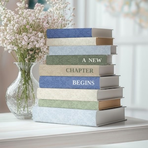 Puede incluir: Una pila de libros con cubiertas decorativas en tonos azules, verdes y beige. El texto "A New Chapter Begins" es visible en los lomos. Un jarrón con flores blancas está a la izquierda.