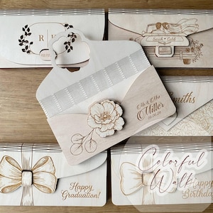 May include: Laser-cut wooden invitation envelopes with various designs. One features a floral design with the initials "R" and "V" and the date "02.02.". Another shows a car with a bride and groom and the words "Just Married". Other envelopes have floral, bow, and text designs.