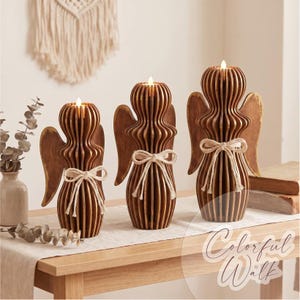 May include: Three brown wooden angel-shaped candle holders of varying heights, each featuring a slatted design, a pair of wings, and a white rope bow tied around the center. Each angel holds a small, lit, battery-operated tea light candle.