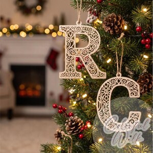 May include: Two light-colored wooden Christmas ornaments shaped like the letters R and C, featuring intricate laser-cut filigree patterns. Each letter hangs from a thin string, suspended among green pine needles, pinecones, and red berries.