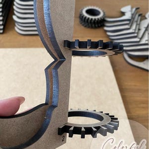 Puede incluir: Un proyecto de artesan&iacute;a en madera parcialmente ensamblado, con una estructura en capas de color marr&oacute;n claro, detalles en negro y componentes en forma de engranaje. El dise&ntilde;o incluye un engranaje circular y el n&uacute;mero "2". Las palabras "Colorful Walk" son visibles.