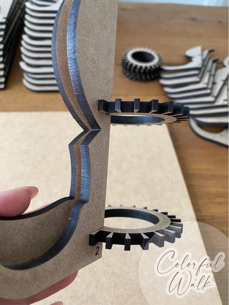 Puede incluir: Un proyecto de artesan&iacute;a en madera parcialmente ensamblado, con una estructura en capas de color marr&oacute;n claro, detalles en negro y componentes en forma de engranaje. El dise&ntilde;o incluye un engranaje circular y el n&uacute;mero "2". Las palabras "Colorful Walk" son visibles.