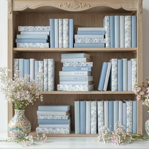 Puede incluir: Una estantería de madera clara llena de libros en tonos azules y blancos. Algunos libros tienen estampados florales, otros son lisos. El texto "ONE FOR THE BOOKS" es visible en los lomos de algunos libros. También hay un jarrón con flores.