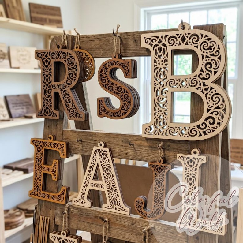 May include: A collection of laser-cut wooden letters featuring intricate filigree patterns, hanging from a rustic wooden ladder display. The letters R, S, B, E, A, J, and I are visible in various wood tones, each suspended by a simple twine loop.