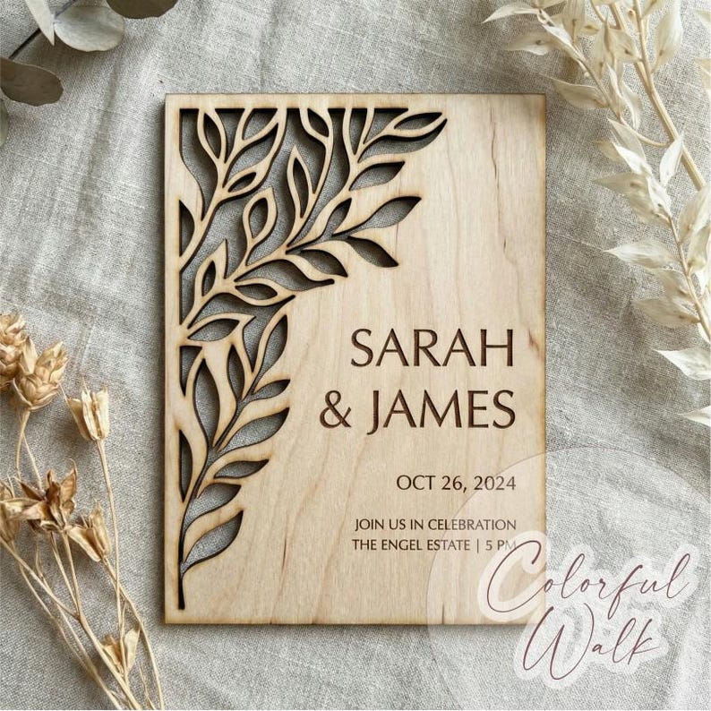 Op de afbeelding: Een houten huwelijksuitnodiging met een lasergesneden bladontwerp in de linkerbovenhoek. De namen "SARAH & JAMES" zijn bruin gedrukt, samen met de datum "26 OKT 2024" en evenementdetails. De uitnodiging is rechthoekig en gemaakt van lichtgekleurd hout.
