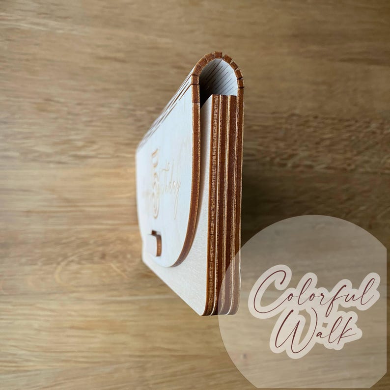 May include: A light-colored wooden box with a curved flap and a small clasp. The text "Happy Birthday" is engraved on the front. The box is made of layered wood and has a natural wood grain.