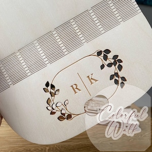 Puede incluir: Una caja de madera de color claro con un dise&ntilde;o decorativo. La caja presenta un patr&oacute;n cortado con l&aacute;ser de l&iacute;neas paralelas y una corona floral con las iniciales "R" y "K". El dise&ntilde;o es de color marr&oacute;n y la caja parece ser de madera.