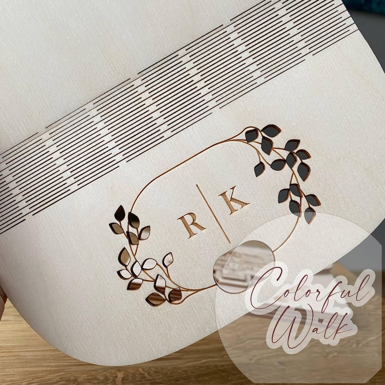 Puede incluir: Una caja de madera de color claro con un dise&ntilde;o decorativo. La caja presenta un patr&oacute;n cortado con l&aacute;ser de l&iacute;neas paralelas y una corona floral con las iniciales "R" y "K". El dise&ntilde;o es de color marr&oacute;n y la caja parece ser de madera.