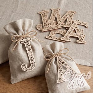 May include: Two light beige linen drawstring pouches, each adorned with a laser-cut wooden letter J or A. Additional laser-cut wooden letters are arranged nearby. The letters feature intricate, swirling filigree patterns throughout.