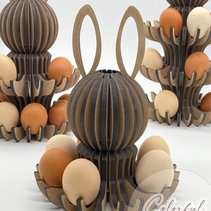 Puede incluir: Soportes de madera para huevos de Pascua con forma de conejos, con m&uacute;ltiples niveles para huevos. Los soportes son marrones y grises, con un dise&ntilde;o de orejas de conejo. Los huevos son de varios tonos de marr&oacute;n y beige. El texto "Colorful Walk" est&aacute; en la esquina inferior derecha.
