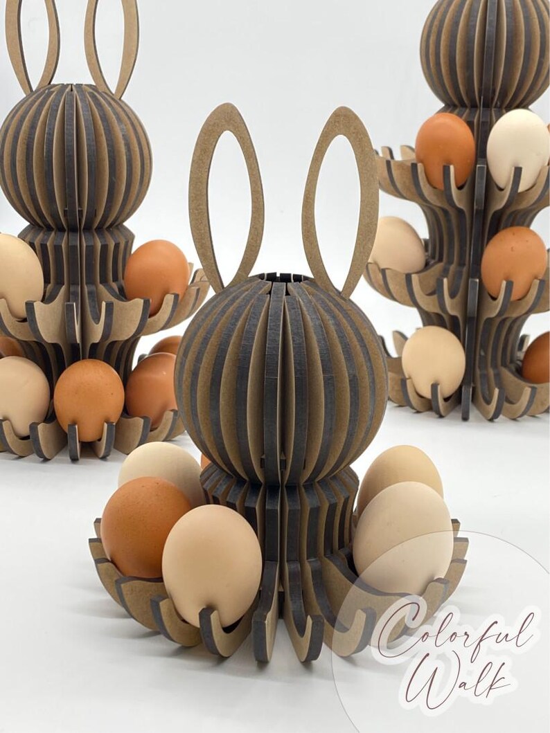 Puede incluir: Soportes de madera para huevos de Pascua con forma de conejos, con m&uacute;ltiples niveles para huevos. Los soportes son marrones y grises, con un dise&ntilde;o de orejas de conejo. Los huevos son de varios tonos de marr&oacute;n y beige. El texto "Colorful Walk" est&aacute; en la esquina inferior derecha.