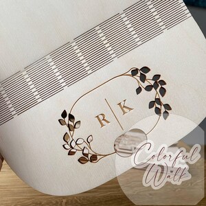 May include: A light-colored wooden box with a decorative design. The box features a laser-cut pattern of parallel lines and a floral wreath with the initials "R" and "K". The design is in a brown color, and the box appears to be made of wood.