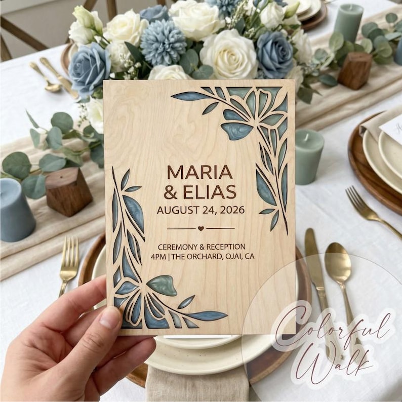 Op de afbeelding: Een houten trouwuitnodiging met lasergesneden bloemmotieven. De uitnodiging bevat de namen "MARIA & ELIAS" en de datum "AUGUST 24, 2026". De uitnodiging wordt in een hand gehouden, met een bloemstuk op de achtergrond.
