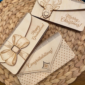May include: Wooden greeting cards with laser-cut designs. One card features a bow, another a deer, and the third a geometric design. The cards have the phrases "Happy Graduation!", "Merry Christmas!", and "Congratulations!" engraved on them.