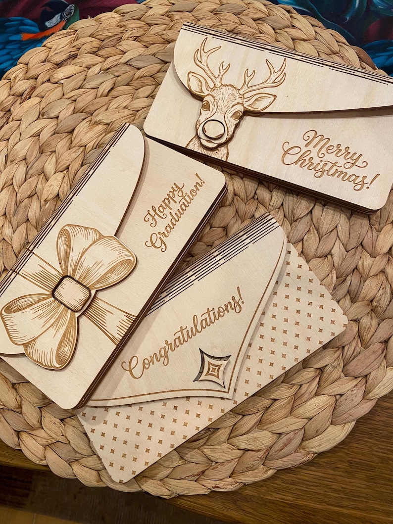 May include: Wooden greeting cards with laser-cut designs. One card features a bow, another a deer, and the third a geometric design. The cards have the phrases "Happy Graduation!", "Merry Christmas!", and "Congratulations!" engraved on them.